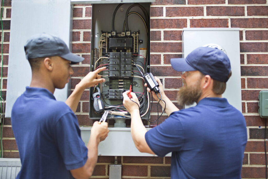 Two men electricians working on a residential breaker box. They use tools to assess the repair and wear matching blue uniforms. The multi-ethnic group is discussing next steps in the job repair. Electric meter to side.